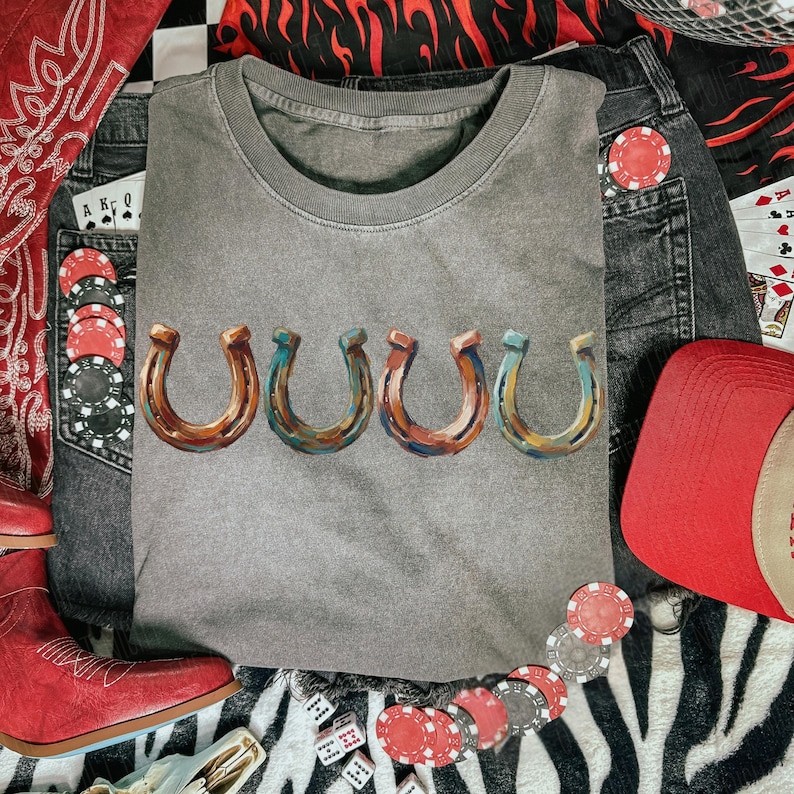 May include: A gray crew neck t-shirt with five horseshoe designs in shades of brown, blue, and orange. The shirt is surrounded by denim shorts, a red baseball cap, poker chips, dice, and a red bandana.