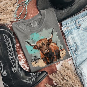 May include: A grey t-shirt with a Highland cow graphic, set against a desert landscape with a turquoise background. The shirt is styled with blue jeans, a black cowboy boot, and a hat, creating a Western-themed ensemble.