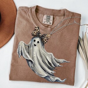 May include: A brown Comfort Colors t-shirt with a smiling ghost graphic and a leopard print bow. A silver necklace with a pendant is draped across the shirt. The shirt has a crew neck and short sleeves. The tag reads "S - P - CH".