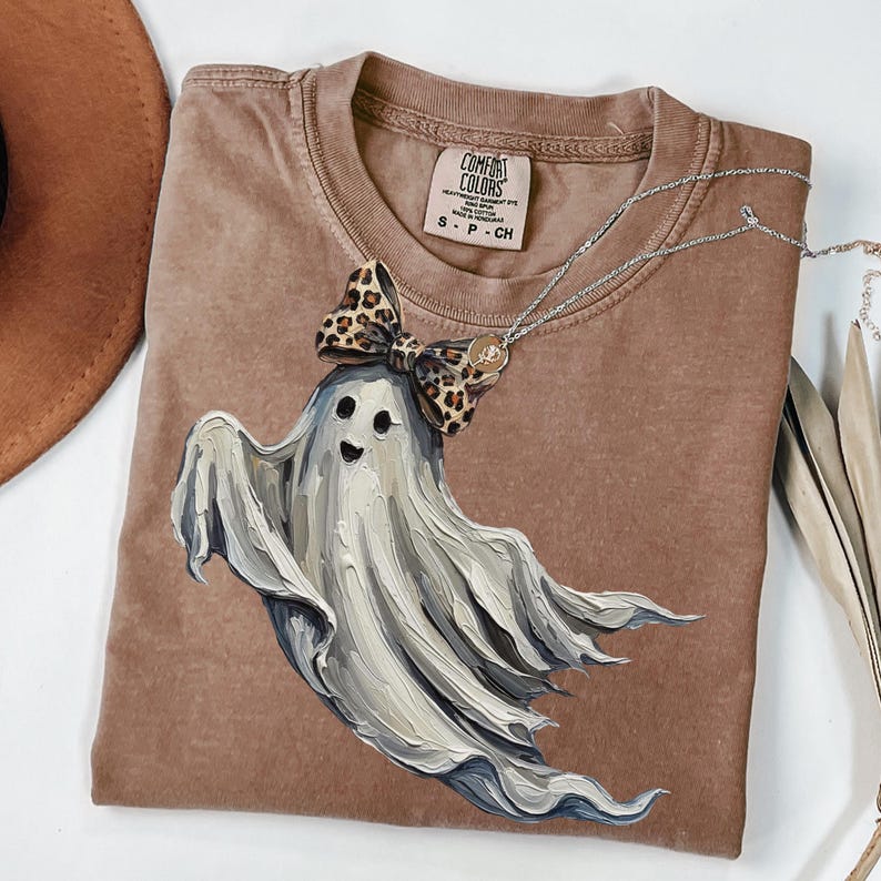 May include: A brown Comfort Colors t-shirt with a smiling ghost graphic and a leopard print bow. A silver necklace with a pendant is draped across the shirt. The shirt has a crew neck and short sleeves. The tag reads "S - P - CH".