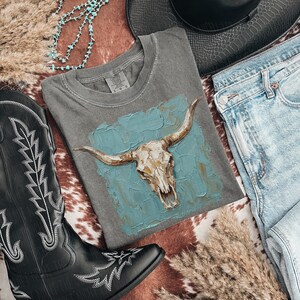 May include: A grey t-shirt with a bull skull design on a turquoise background. The shirt is styled with light-wash denim jeans, a black cowboy hat, and black cowboy boots. A turquoise necklace and dried grass complete the western theme.