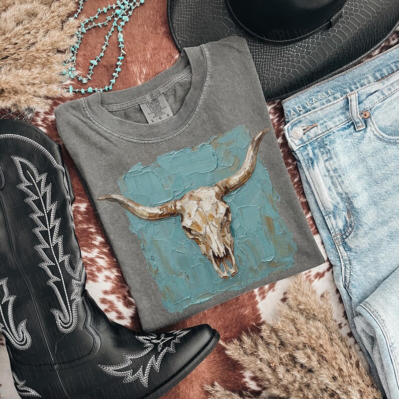 May include: A grey t-shirt with a bull skull design on a turquoise background. The shirt is styled with light-wash denim jeans, a black cowboy hat, and black cowboy boots. A turquoise necklace and dried grass complete the western theme.