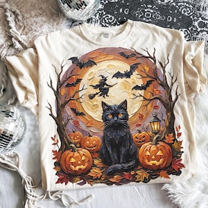 May include: A cream-colored t-shirt featuring a graphic print of a black cat sitting in front of a large full moon. The scene includes orange jack-o'-lanterns, bare trees, flying bats, a witch on a broomstick, and a small lantern on the ground.