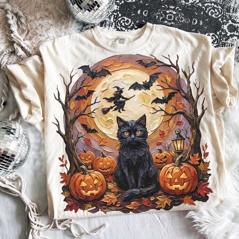 May include: A cream-colored t-shirt featuring a graphic print of a black cat sitting in front of a large full moon. The scene includes orange jack-o'-lanterns, bare trees, flying bats, a witch on a broomstick, and a small lantern on the ground.