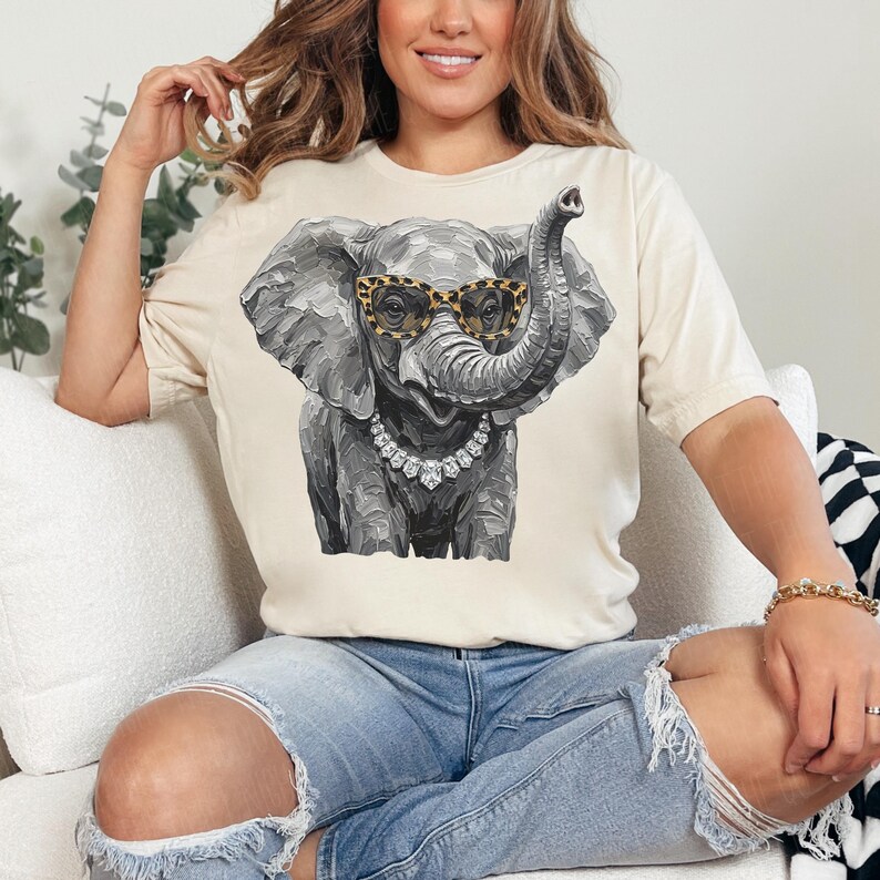 May include: A cream-colored short-sleeve t-shirt featuring a graphic illustration of a gray elephant wearing leopard-print glasses and a silver-toned crystal necklace. The elephant is depicted with a textured, painterly style and its trunk is raised.