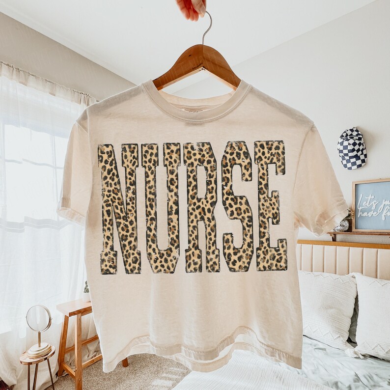 May include: A cream-colored t-shirt with the word "NURSE" printed in large, bold letters. The letters are filled with a leopard print pattern in shades of brown and tan. The shirt is hanging on a wooden hanger.
