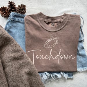 May include: A brown t-shirt with a white football graphic and the word "Touchdown" in a script font. The shirt is paired with blue denim shorts and a brown sweater. Decorative pinecones are in the background.