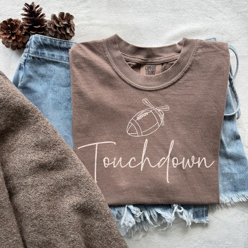 May include: A brown t-shirt with a white football graphic and the word "Touchdown" in a script font. The shirt is paired with blue denim shorts and a brown sweater. Decorative pinecones are in the background.