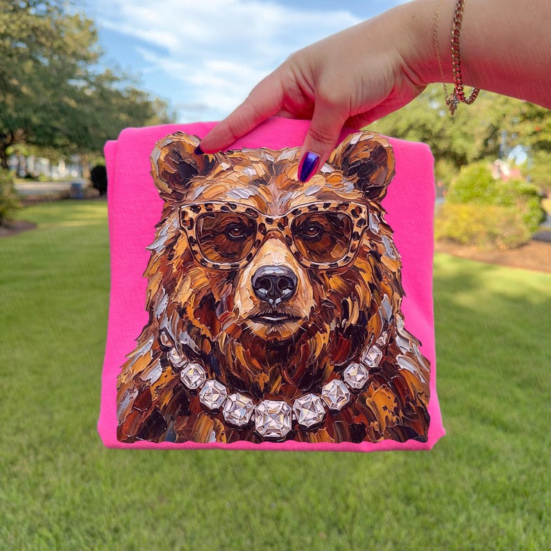 May include: A hot pink t-shirt featuring a detailed, artistic rendering of a bear's face. The bear wears leopard print glasses and a diamond necklace. The bear's fur is rendered in shades of brown, tan, and gold.