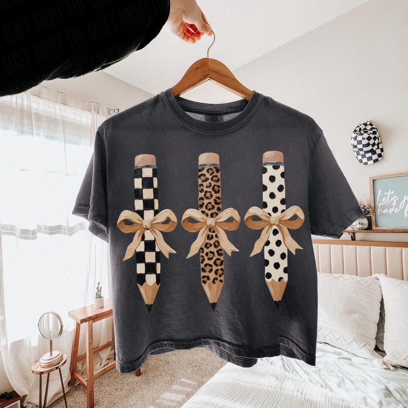 以下が含まれることがあります： 3つの鉛筆のデザインとリボンが付いた濃い灰色のTシャツ。鉛筆は、白黒チェック、ヒョウ柄、白地に黒い水玉模様の異なるパターンです。リボンはベージュ色です。