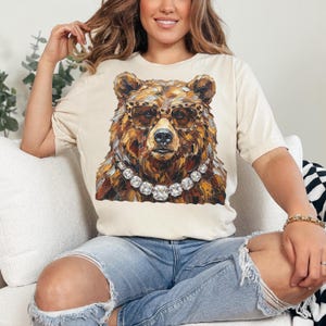 May include: Cream-colored t-shirt featuring a detailed illustration of a bear wearing leopard print glasses and a diamond necklace. The bear's fur is rendered in shades of brown and gold. The shirt has short sleeves and a relaxed fit.