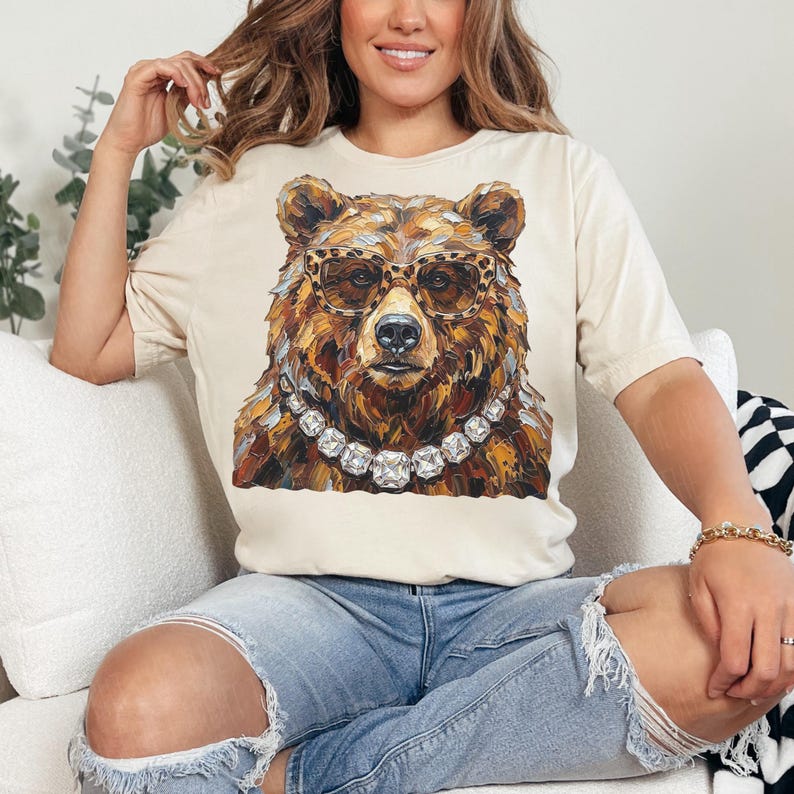 May include: Cream-colored t-shirt featuring a detailed illustration of a bear wearing leopard print glasses and a diamond necklace. The bear's fur is rendered in shades of brown and gold. The shirt has short sleeves and a relaxed fit.