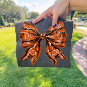 May include: A gray tote bag featuring a large, hand-painted bow design. The bow is primarily orange with black and white accents, resembling a basketball court. The bag is held against a backdrop of green grass and a blue sky.