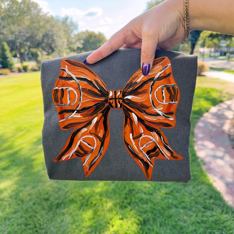 May include: A gray tote bag featuring a large, hand-painted bow design. The bow is primarily orange with black and white accents, resembling a basketball court. The bag is held against a backdrop of green grass and a blue sky.