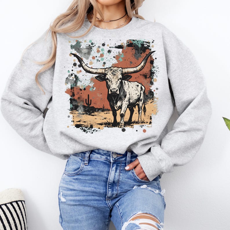 May include: A light gray sweatshirt featuring a graphic of a longhorn steer with large horns. The steer is set against a backdrop of a desert landscape with a color splash design in shades of brown, black, and teal.