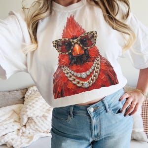 May include: White t-shirt featuring a detailed illustration of a red cardinal wearing leopard print sunglasses and a gold chain necklace with diamond accents. The bird has a serious expression. The shirt is paired with blue jeans.