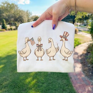 May include: White sweatshirt featuring four cartoon geese. One goose holds an American flag, another has a bow tie, a third holds a sparkler, and the last wears a patriotic top hat. The geese are in a row.