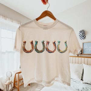 May include: Cream-colored t-shirt featuring five horseshoe designs in shades of brown, teal, and copper. The horseshoes are arranged horizontally across the front of the shirt. The shirt is hanging on a wooden hanger.
