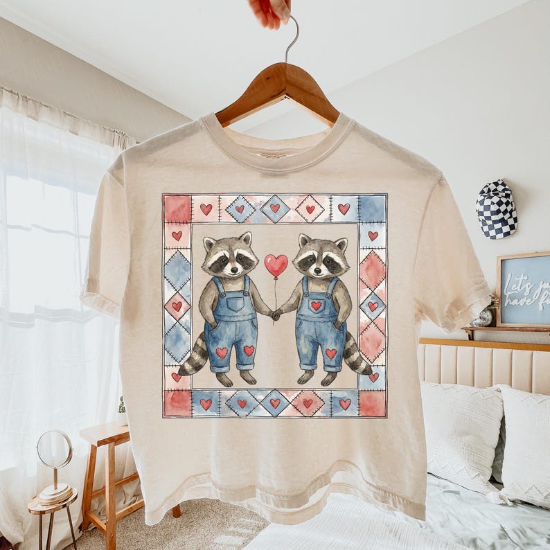 Pu&ograve; includere: T-shirt color crema con un'illustrazione ad acquerello di due procioni che si tengono per mano e un palloncino a forma di cuore. I procioni indossano salopette blu e sono circondati da un bordo quadrato con cuori e motivi a rombi.