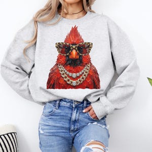 May include: A light gray sweatshirt featuring a vibrant red cardinal wearing leopard print sunglasses and a gold chain necklace. The bird is the focal point, creating a bold and stylish statement.