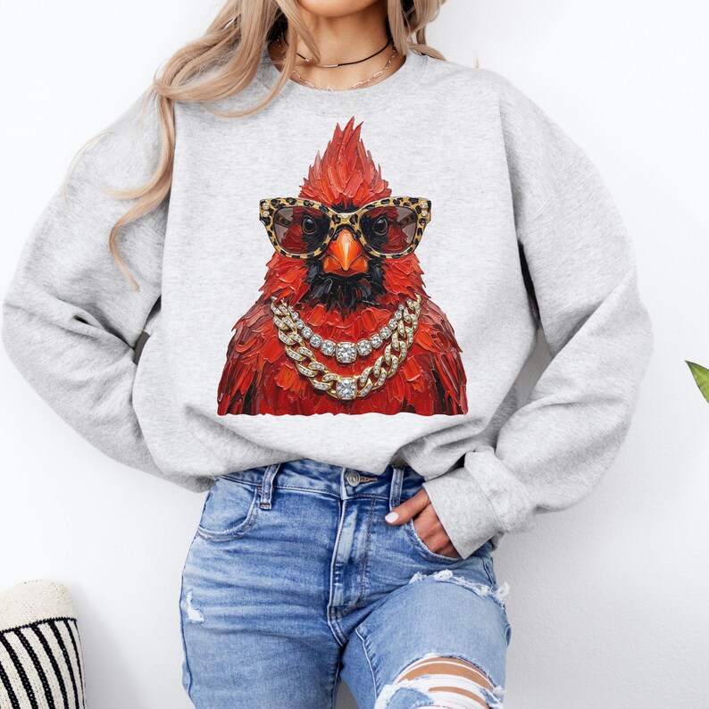 May include: A light gray sweatshirt featuring a vibrant red cardinal wearing leopard print sunglasses and a gold chain necklace. The bird is the focal point, creating a bold and stylish statement.