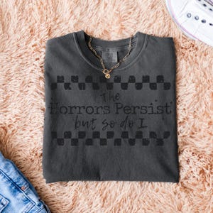 May include: Dark gray t-shirt with the text "The Horrors Persist but so do I" in a distressed font. The shirt has a checkerboard design above and below the text. A gold necklace with a pendant rests on the shirt.