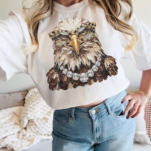 May include: White t-shirt featuring a detailed illustration of an eagle wearing leopard print glasses and a diamond necklace. The eagle's feathers are rendered in shades of brown, beige, and white. The shirt is a casual, everyday wear item.