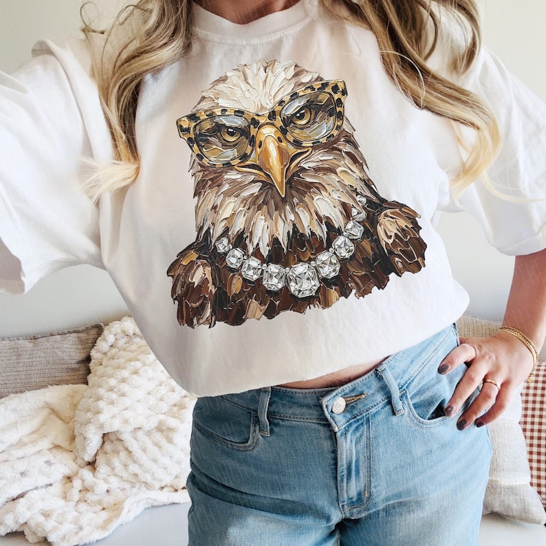 May include: White t-shirt featuring a detailed illustration of an eagle wearing leopard print glasses and a diamond necklace. The eagle's feathers are rendered in shades of brown, beige, and white. The shirt is a casual, everyday wear item.