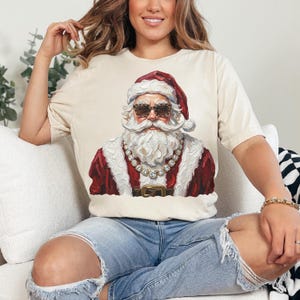 May include: A cream-colored short-sleeve t-shirt featuring a graphic print of Santa Claus wearing leopard-print sunglasses and a thick gold chain necklace. The illustration shows Santa with a white beard, a red hat, and a red coat with white trim.