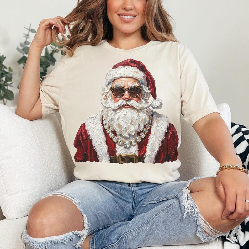 May include: A cream-colored short-sleeve t-shirt featuring a graphic print of Santa Claus wearing leopard-print sunglasses and a thick gold chain necklace. The illustration shows Santa with a white beard, a red hat, and a red coat with white trim.