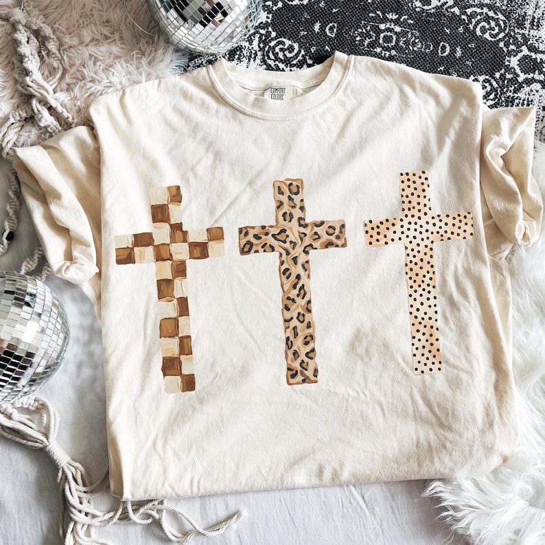 May include: Cream-colored t-shirt featuring three crosses with unique patterns. One cross has a checkerboard design, another a leopard print, and the third has a polka dot pattern. The shirt is made of a soft material.