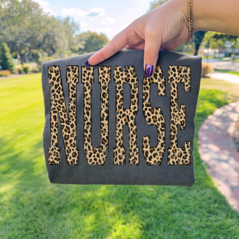 May include: A dark gray t-shirt with the word "NURSE" in large, bold letters. The letters are filled with a leopard print pattern. The t-shirt is held up against a background of green grass and trees.
