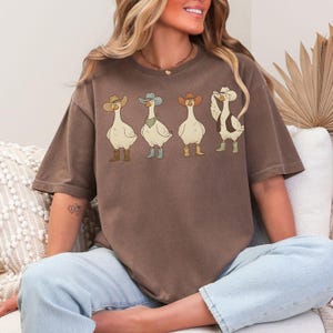 May include: Brown t-shirt featuring four cartoon geese wearing cowboy hats and boots. The geese are in various poses, with one adjusting its hat. The shirt is a relaxed fit and has a vintage look.
