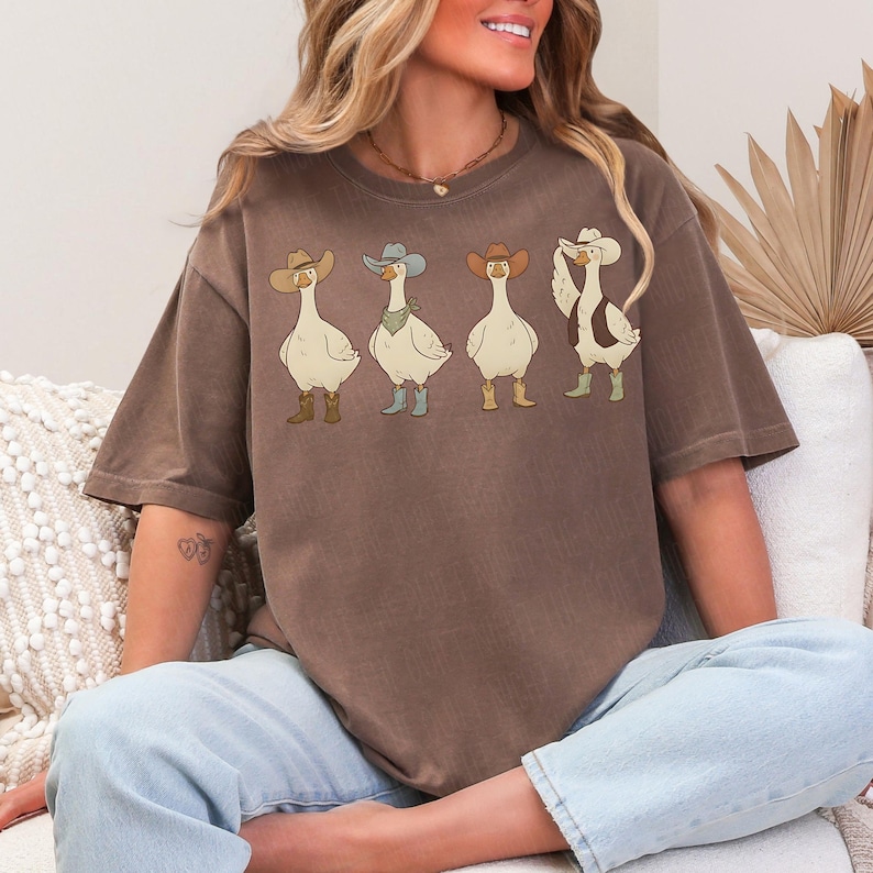 May include: Brown t-shirt featuring four cartoon geese wearing cowboy hats and boots. The geese are in various poses, with one adjusting its hat. The shirt is a relaxed fit and has a vintage look.