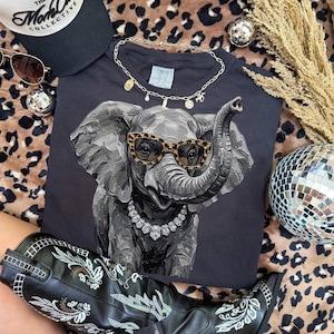 May include: A dark gray t-shirt featuring a textured, painterly illustration of an elephant wearing leopard-print sunglasses and a silver-toned crystal necklace. The elephant is depicted with visible brushstrokes in shades of gray and black.