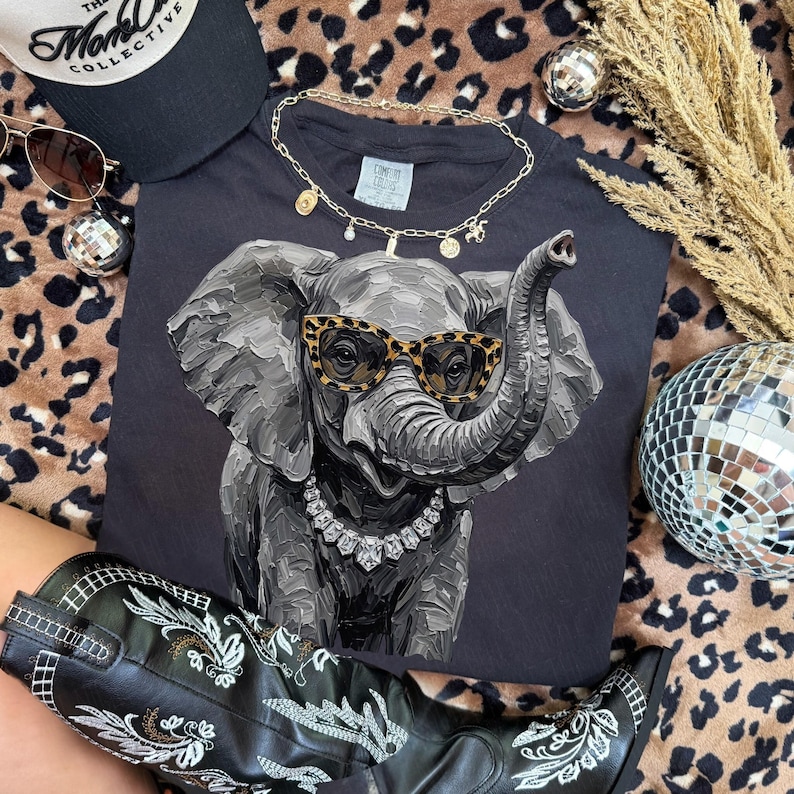 May include: A dark gray t-shirt featuring a textured, painterly illustration of an elephant wearing leopard-print sunglasses and a silver-toned crystal necklace. The elephant is depicted with visible brushstrokes in shades of gray and black.