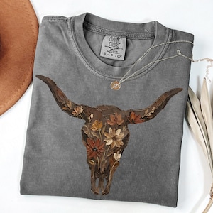 May include: A gray Comfort Colors t-shirt featuring a detailed illustration of a bull skull adorned with flowers in shades of brown, orange, and cream. A delicate silver necklace with a small pendant rests on the shirt. The shirt has a small tag that says S-P-CH.