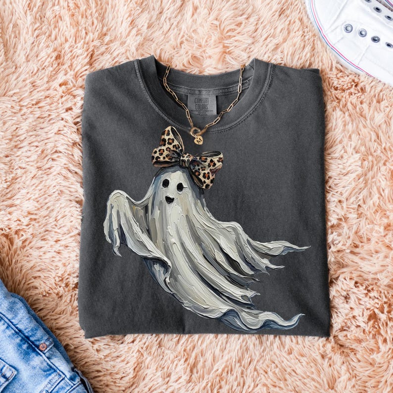 May include: Dark grey t-shirt with a ghost illustration and a leopard print bow. A gold chain necklace is on the shirt. The t-shirt is folded on a pink, fluffy surface.