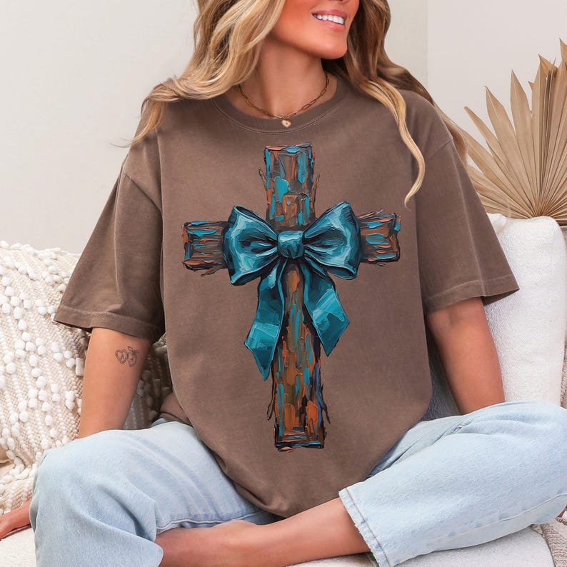 Peut inclure: Un t-shirt marron avec un motif de croix et un n&oelig;ud bleu sarcelle. La croix a un aspect rustique et textur&eacute; avec des nuances de marron, d'orange et de bleu sarcelle. Le t-shirt est de coupe d&eacute;contract&eacute;e, avec un jean bleu clair.