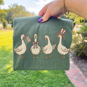 May include: Green t-shirt featuring a whimsical illustration of four geese. One goose holds an American flag, another wears a bow tie, and the last wears a patriotic Uncle Sam hat. The geese are in a row.