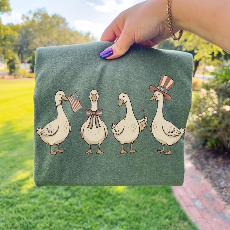 May include: Green t-shirt featuring a whimsical illustration of four geese. One goose holds an American flag, another wears a bow tie, and the last wears a patriotic Uncle Sam hat. The geese are in a row.
