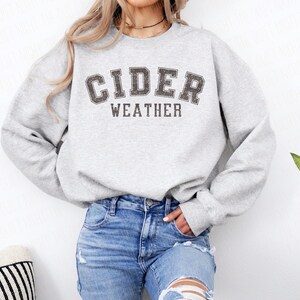 K&ouml;nnte beinhalten: Hellgraues Sweatshirt mit dem Aufdruck "CIDER WEATHER" in verblichener, dunkelgrauer Schrift. Das Sweatshirt wird mit einer blauen Jeans kombiniert. Der Stil ist l&auml;ssig und bequem.