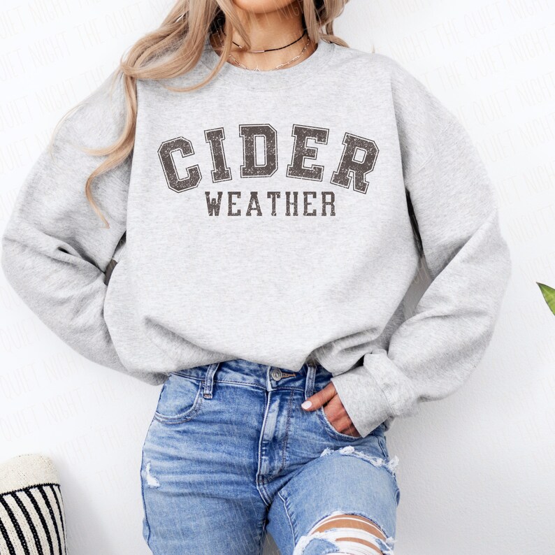 K&ouml;nnte beinhalten: Hellgraues Sweatshirt mit dem Aufdruck "CIDER WEATHER" in verblichener, dunkelgrauer Schrift. Das Sweatshirt wird mit einer blauen Jeans kombiniert. Der Stil ist l&auml;ssig und bequem.