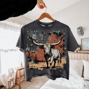 May include: Dark gray t-shirt featuring a longhorn illustration with a desert landscape backdrop. The design includes a bull with large horns, a cactus, and colorful paint splatters. The shirt is hanging on a wooden hanger.