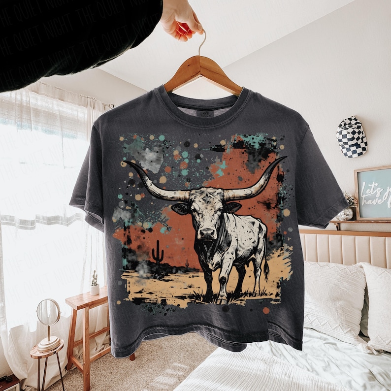 May include: Dark gray t-shirt featuring a longhorn illustration with a desert landscape backdrop. The design includes a bull with large horns, a cactus, and colorful paint splatters. The shirt is hanging on a wooden hanger.