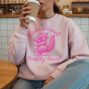 May include: A light pink sweatshirt with a pink raccoon graphic and the text "Kinda Sweet Mostly Feral". The raccoon is standing on its hind legs. The person is holding a brown paper coffee cup.