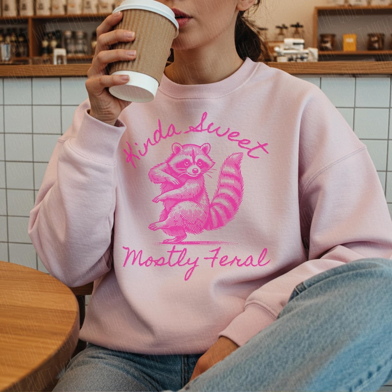 May include: A light pink sweatshirt with a pink raccoon graphic and the text "Kinda Sweet Mostly Feral". The raccoon is standing on its hind legs. The person is holding a brown paper coffee cup.
