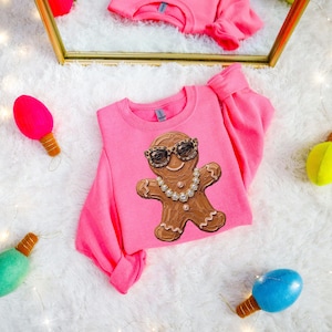 May include: A bright pink long-sleeve sweatshirt featuring a central graphic of a brown gingerbread person wearing leopard print sunglasses and a silver-toned rhinestone necklace. The sweatshirt has a crew neckline and ribbed cuffs.