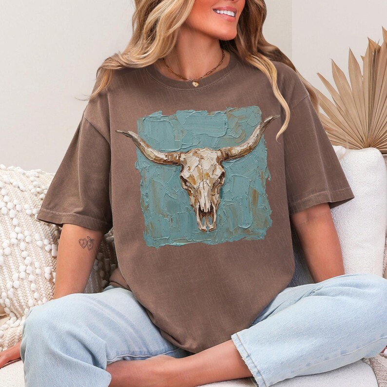 May include: Brown t-shirt with a longhorn skull design on a teal blue textured background. The skull is detailed with tan and cream colours, creating a rustic, western aesthetic. The shirt is a relaxed fit.