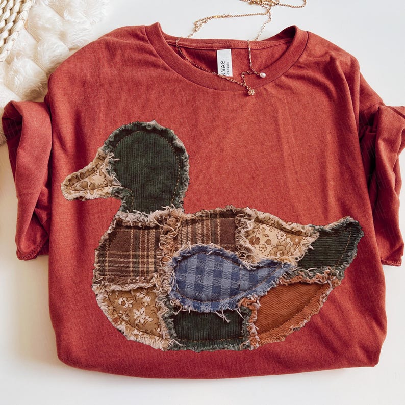 May include: A rust-colored crew neck t-shirt featuring a large, central duck-shaped appliqu&eacute;. The duck is constructed from various fabric patches, including corduroy, plaid, and floral patterns, with raw, frayed edges stitched onto the shirt.